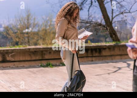 Hübsches Schulmädchen, das ein Buch liest, während es auf dem Schulhof spaziert. Sie trägt die Schultasche an der anderen Hand Stockfoto