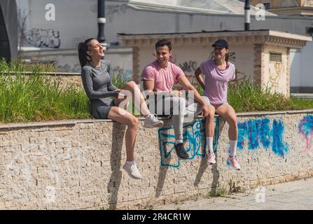 Drei Sportfreunde, die sich vom Workout erholen. Sie sitzen auf einem Wandvorsprung, unterhalten sich und lachen Stockfoto