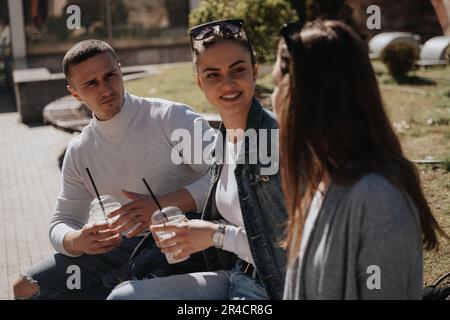 Drei beste Freunde, die sich unterhalten, während sie im Park sitzen und Milchshake-Tassen halten Stockfoto