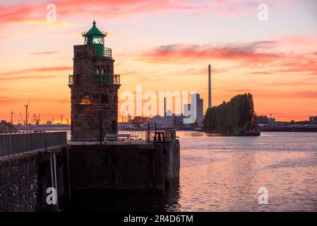 Historischer Leuchtturm am Flusshafen bei Sonnenuntergang. Industriegebäude und Windturbinen sind in der Ferne sichtbar Stockfoto