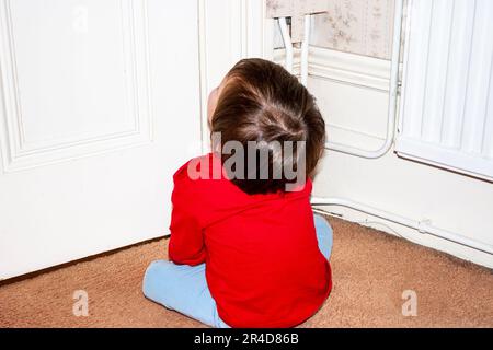 Seltene Aussicht auf ein zweijähriges Kind, Junge, das vor einer weißen Innentür sitzt und erwartungsvoll nach oben blickt. Stockfoto