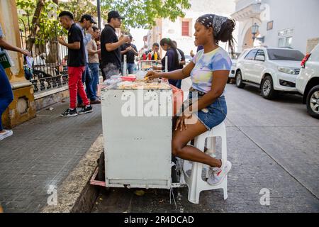 Eine junge Frau sitzt an einem Areas Street Food Stand, während die Leute in der Nähe beim Essen stehen Stockfoto