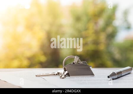 Nahaufnahme eines hausförmigen Schlüsselrings mit Schlüsseln und einem Stift auf einem Kaufvertrag für ein Haus auf einem Tisch vor einem Fenster mit Bäumen auf der Straße. Stockfoto