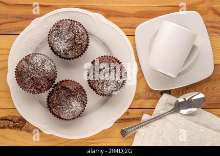 Vier hausgemachte Schokoladen-Cupcakes mit weißen Keramikgerichten auf einem Holztisch, Makro, Draufsicht. Stockfoto