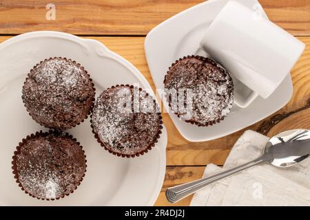 Vier hausgemachte Schokoladen-Cupcakes mit weißen Keramikgerichten auf einem Holztisch, Makro, Draufsicht. Stockfoto