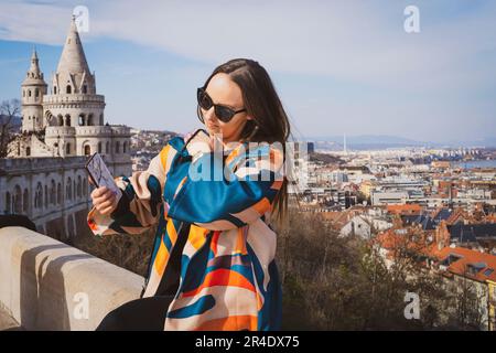 Eine junge Frau in einer Sonnenbrille mit einem Telefon in der Hand in einer bunten Jacke in Budapest Stockfoto