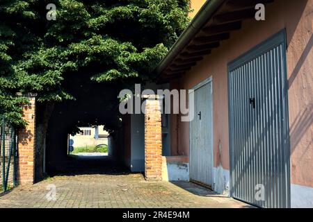 Gepflasterter Pfad teilweise im Schatten unter Bäumen im Park eines Dorfes an einem sonnigen Tag Stockfoto