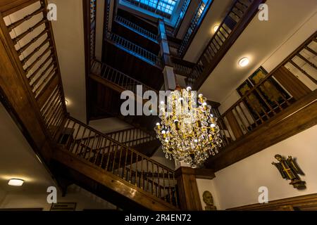 Das ehemalige Hotel Münchnerhof in der Dreifaltigkeitsgasse 3, Salzburg, Österreich, verfügt über 100 Zimmer, die über eine Treppe mit dunkler Holztäfelung, die von einem Glasdach und einem riesigen Kristallleuchter beleuchtet ist, erreicht werden. Man kann dort noch viele geschnitzte Figuren mit dem Münchner Kindl-Motiv sehen Stockfoto