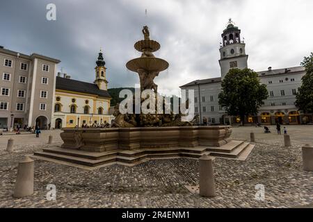 Die Residenzfontäne mit ihren Tritonen- und Pferdefiguren steht vor dem Hintergrund des Glockenturms und einer Kirche auf dem Residenzplatz in Salzburg, Österreich. Fürsterzbischof Guidobald Graf von Thun und Hohenstein ließ die 15 Meter hohe barocke Fontäne, gehauen aus Untersberger Marmor, zwischen 1656 und 1661 errichten. Stockfoto