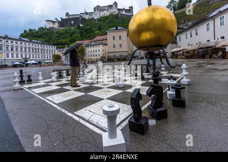 Schachspieler auf dem Kapitelplatz in Salzburg (Osterreich). Im Hintergrund die Festung Hohensalzburg. Die Sphaera-Sphäre wurde vom deutschen Künstler Stephan Balkenhol geschaffen. Auf einer goldenen Kugel mit einem Durchmesser von 5 Metern steht eine männliche Figur aus Bronze mit schwarzer Hose und weißem Hemd Stockfoto