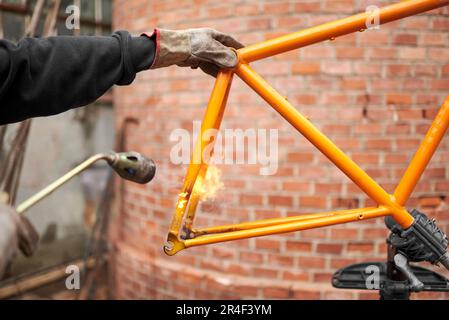 Eine nicht wiedererkennbare Person, die mit einem Lötbrenner die Farbe eines orangefarbenen Fahrradrahmens entfernt, der gerade in seiner Werkstatt renoviert wird. Stockfoto