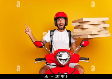 Ein bärtiger Liefermann mit rotem Helm und weißem T-Shirt sitzt auf einem roten Roller mit einem Haufen Pizzaschachteln in einer Hand und zeigt mit dem Zeigefinger auf einen leeren Raum Stockfoto