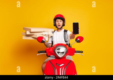 Ein junger Mann mit rotem Helm und weißem T-Shirt sitzt auf einem roten Roller mit einem Haufen Pizzaschachteln in einer Hand, zeigt Telefonbildschirm, schnelle Lieferung Stockfoto