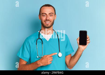 Junger männlicher Mediziner hält den Telefonbildschirm in der Hand und zeigt auf ihn, schaut auf die Kamera und lächelt, trägt ein spezielles medizinisches Hemd, professionelle Leute Stockfoto