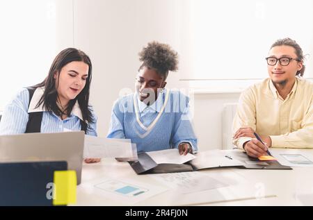 Ein vielseitiges Team von Kollegen, darunter eine Chinesin, eine Afroamerikanerin und eine Kaukasierin, die Ideen an einem Schreibtisch diskutieren. Die weiße Frau hat Papiere Stockfoto