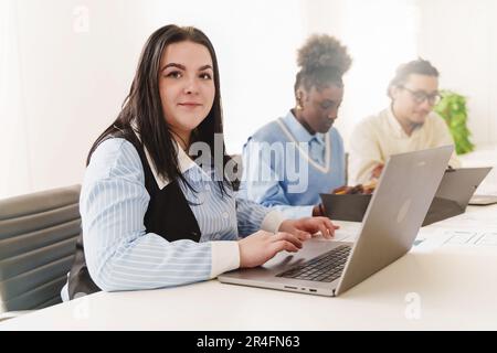 Weiße Frau mit Piercing, die auf einem Laptop arbeitet – in einem vielfältigen Büro konzentriert sich eine junge Frau auf ihren Laptop, ihre afrikanischen und chinesischen Kollegen verschwommen Stockfoto
