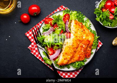 Gebratenes weißes Fischfilet mit Gemüsesalat aus Salat, Kirschtomaten und roten Zwiebeln, schwarzer Tischhintergrund, Draufsicht Stockfoto