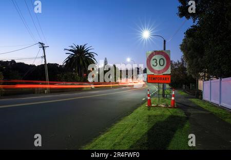 Temporäres Schild mit 30km Geschwindigkeitsbegrenzung und Verkehrskegel am Straßenrand. Autolichtspuren an der Kreuzung. Auckland. Stockfoto