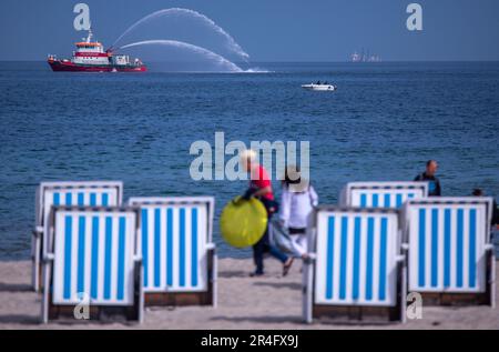 Rostock, Deutschland. 28. Mai 2023. Das Feuerboot 'Albert Wegener' spritzt Wasser, während die ersten Urlauber morgens an den meist noch leeren Liegen an der Ostseeküste vorbeilaufen. Mit viel Sonnenschein und hohen Temperaturen zeigt das Wetter seine beste Seite am Pfingstsonntag in Norddeutschland. Kredit: Jens Büttner/dpa/Alamy Live News Stockfoto