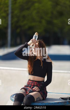 Erschöpfte Skaterin, die in einem Betonpool sitzt und eine Glasflasche kaltes Wasser in der Hand hält. Coole junge Erwachsene Rollblader, die sich in einem Skatepark entspannen Stockfoto