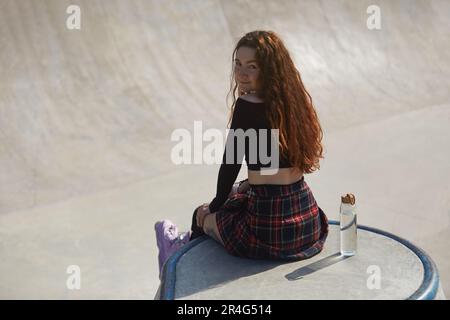 Junge Inline-Rollschuh-Frau, die sich in einem Eislaufbecken posiert. Porträt eines Skaters, der mit einer Glasflasche Wasser auf einer Rampe sitzt. Stockfoto