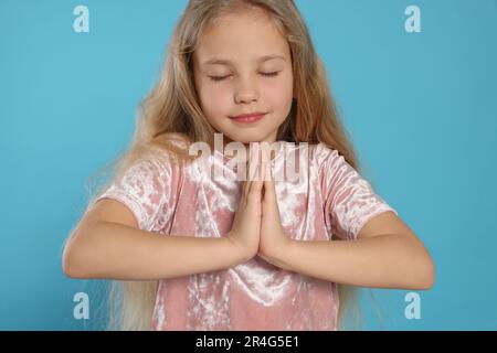 Hübsches Mädchen mit aneinander gefalteten Händen und geschlossenen Augen, betend auf hellblauem Hintergrund Stockfoto