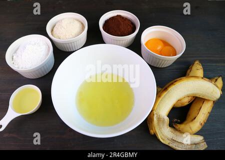Eiweiß in einer Rührschüssel, umgeben von Zutaten für den Backen von Bananenschokolade Stockfoto