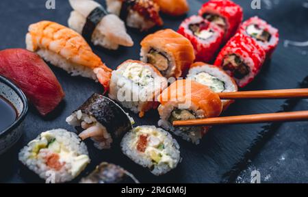 Farbenfrohes Sushi auf schwarzem Schreibtisch mit California Roll. Eine Person nimmt Maki mit Stäbchen. Orientalische japanische gesunde Küche mit rohem Fisch und Reis Stockfoto