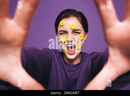 Seltsame junge Frau, die ein Selfie mit lächelnden Aufklebern im Gesicht macht. Verspielte Frau, die fröhlich lächelt, während sie ein Foto von sich selbst macht. Fröhliches junges w Stockfoto