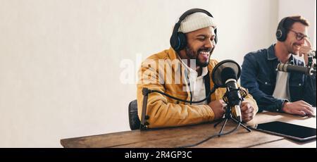 Ein Radio-dj lächelt, während er in einem Studio in ein Mikrofon spricht. Glücklicher junger Mann, der zusammen mit einem Gast eine Audioübertragung moderiert. Zwei junge Inhalte c Stockfoto