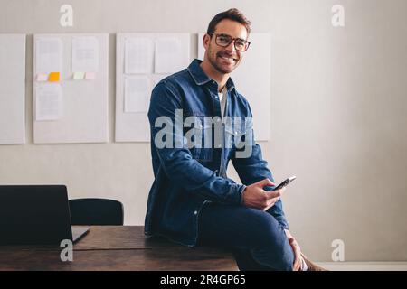 Glücklicher junger Geschäftsmann, der in die Kamera lächelt, während er mit einem Smartphone auf dem Schreibtisch sitzt. Fröhlicher Geschäftsmann mit Brille, der aus der Ferne in einem cr arbeitet Stockfoto