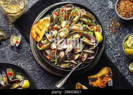 Meeresfrüchtesuppe mit köstlichen Muscheln in schwarzer Schüssel garniert mit frischen Kräutern und rotem Pfeffer. Serviert mit Zitrone, knusprigem Brot und Getränk auf dunklem Hintergrund. Stockfoto