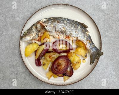 Meeresfrüchte, Blick von oben köstliche gegrillte Seebarschfische auf Teller, serviert mit Kartoffeln und Zwiebeln auf grauem Hintergrund oder Oberfläche mit selektivem Fokus. Heidenmeer Stockfoto