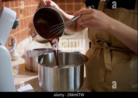 Nahaufnahme-Konditor, der geschmolzene Schokolade in eine Planetenmischschüssel gießt und diese mit Sahne oder Eiweiß mischt Stockfoto