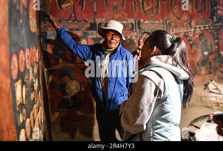 (230528) -- ZANDA, 28. Mai 2023 (Xinhua) -- Rigzin Wangzhab stellt Touristen Wandbilder in einer Grotte in der Präfektur Zanda County der Präfektur Ngari, Südwestchina, Autonome Region Tibet, vor, 26. Mai 2023. Versteckt zwischen Sandsteinhügeln im abgelegenen Westen Tibets liegt ein Abschnitt von honigwabenförmigen Höhlen weit außerhalb der Reichweite der meisten Reisenden. Die 1.000 Jahre alten Höhlen in der Präfektur Ngari in der Autonomen Region Tibet, die als Donggar- und Piyang-Grotten bekannt sind, beherbergen eine der weltweit größten Sammlungen tibetischer buddhistischer Wandbilder. Rigzin Wangzhab, 75, bewacht das Gelände seit über 20 Jahren. Stockfoto