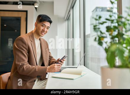 Ein junger, lächelnder japanischer Geschäftsmann, der im Büro ein Handy benutzt. Stockfoto