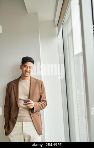 Glücklicher junger asiatischer Geschäftsmann mit Smartphone im Büro, vertikal. Stockfoto