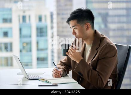 Junger asiatischer Geschäftsmann, der an einem Laptop arbeitet, im Büro Kaffee trinkt. Stockfoto