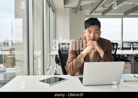 Ein junger asiatischer Geschäftsmann, der im Büro mit einem Laptop in die Kamera schaut. Stockfoto