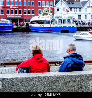 Stavanger, Rogaland, Norwegen, 19 2023. Mai, mittelalterliches Paar, das entspannt am Ufer des Stavanger Hafens sitzt, mit traditionellen alten Hafengebäuden und Stockfoto