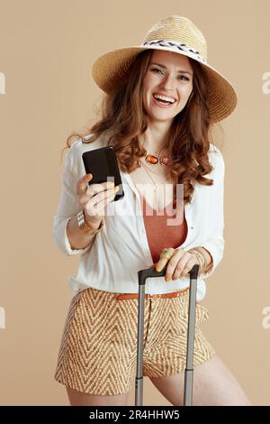 Strandurlaub. Lächelnde elegante Hausfrau in weißer Bluse und Shorts isoliert auf beigem Hintergrund mit Strohhut und Trolley-Tasche mit Smartphone. Stockfoto