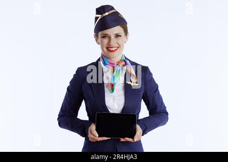 Glückliche, moderne Stewardess mit weißem Hintergrund in Uniform, die den leeren Bildschirm des Tablet-PCs zeigt. Stockfoto