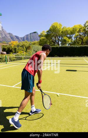 Seitenansicht eines birassischen jungen Mannes, der Tennisball mit Schläger auf dem Tennisplatz vor klarem Himmel bedient Stockfoto