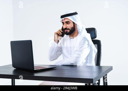 Emirat, mittelöstlicher Geschäftsmann mit Kandora, der im Büro arbeitet, Dubai - ein attraktiver Geschäftsmann, der am Computer-Schreibtisch und an der Fernbedienung sitzt Stockfoto