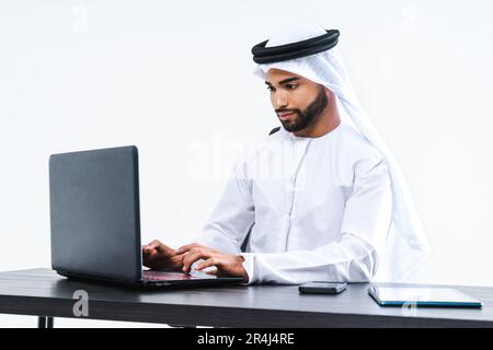Emirat, mittelöstlicher Geschäftsmann mit Kandora, der im Büro arbeitet, Dubai - ein attraktiver Geschäftsmann, der am Computer-Schreibtisch und an der Fernbedienung sitzt Stockfoto