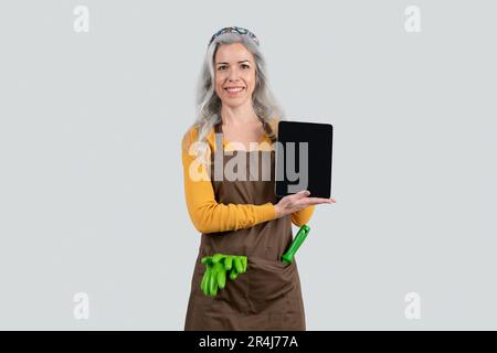 Eine glückliche alte, grauhaarige frau in Schürze mit Gummihandschuhen zeigt ein Tablet mit leerem Bildschirm Stockfoto
