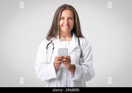 Porträt einer fröhlichen Therapeutin in weißem Kittel und mit Stethoskop mit Mobiltelefon für die Arbeit, grauer Hintergrund Stockfoto