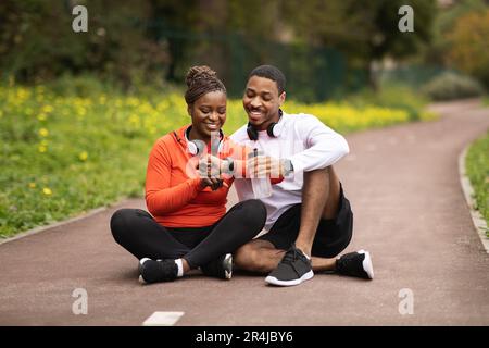 Die positive junge schwarze Familie in Sportbekleidung sitzt auf der Straße und ruht sich mit Flaschen Wasser aus. Überprüfe den Puls auf dem FIT-Tracker Stockfoto