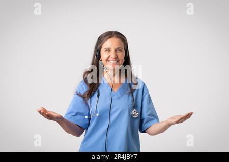 Glückliche Seniorin in weißem Mantel mit Stethoskop in Kopfhörern, beratende Patientin, die auf grauem Hintergrund posiert Stockfoto