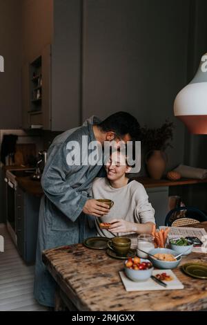 Ein Mann küsst eine lächelnde nicht binäre Person, während er zu Hause frühstückt Stockfoto